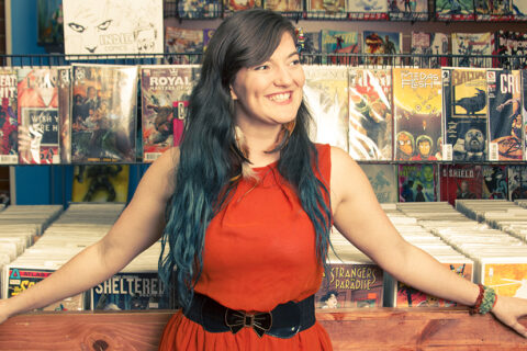 Woman in red dress leans on comic book stand while she smiles